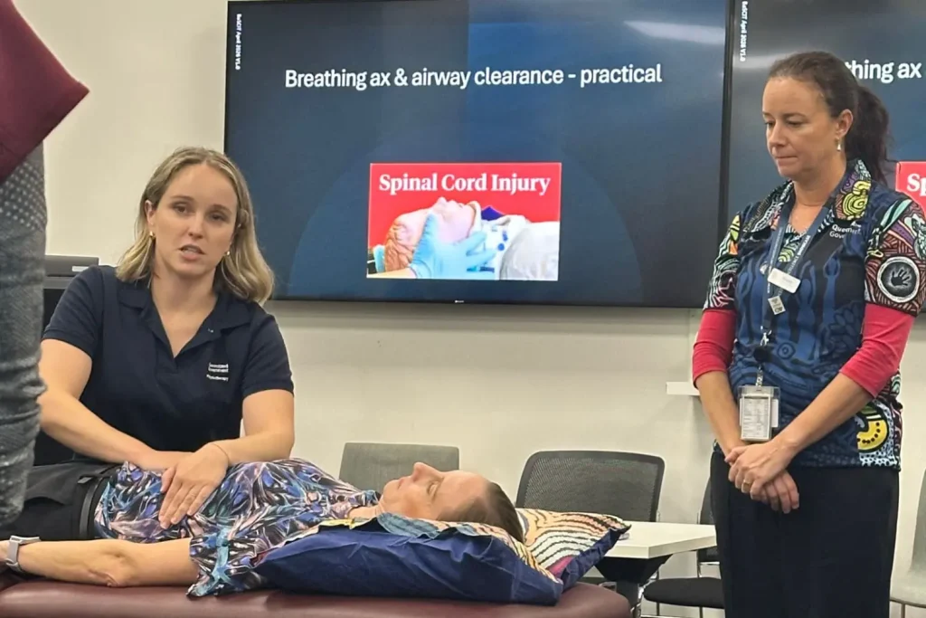 A photograph of a medical training session focused on breathing and airway clearance in spinal cord injury patients. Two instructors demonstrate practical techniques on a person lying on a table, with a screen displaying the title "Breathing ax & airway clearance - practical" and an image labelled "Spinal Cord Injury" in the background.