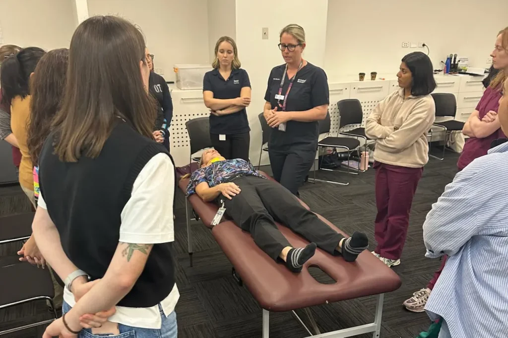 A photograph showing a group of people standing around a person lying on a medical examination table during a training or demonstration session. The setting appears to be a classroom or clinical environment with chairs, medical equipment, and individuals attentively observing or participating in the activity.