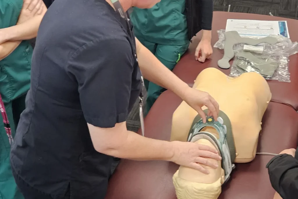 A photograph of a medical training session focused on breathing and airway clearance in spinal cord injury patients. Two instructors demonstrate practical techniques on a person lying on a table, with a screen displaying the title "Breathing ax & airway clearance - practical" and an image labelled "Spinal Cord Injury" in the background.