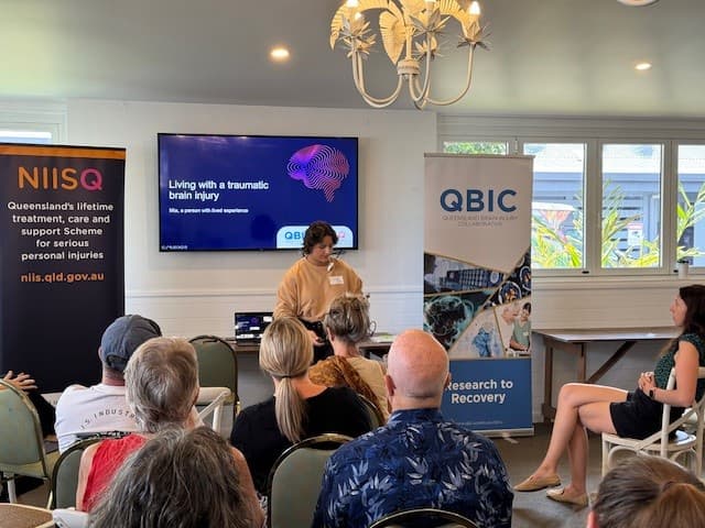 A young woman with short hair addresses a room of seated people. Behind her a presentation titled, "living with a traumatic brain injury" is visible. either side are banners that read "NIISQ" and "QBIC".