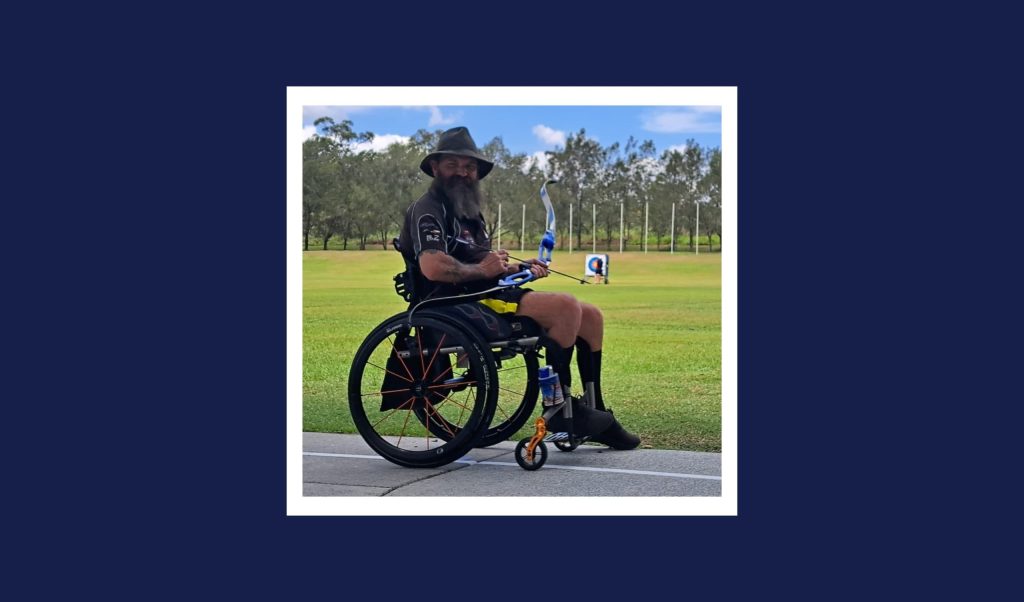A man with a beard and hat in a wheel chair holds a bow and arrow. Behind him, archery targets, grass and trees are visible.