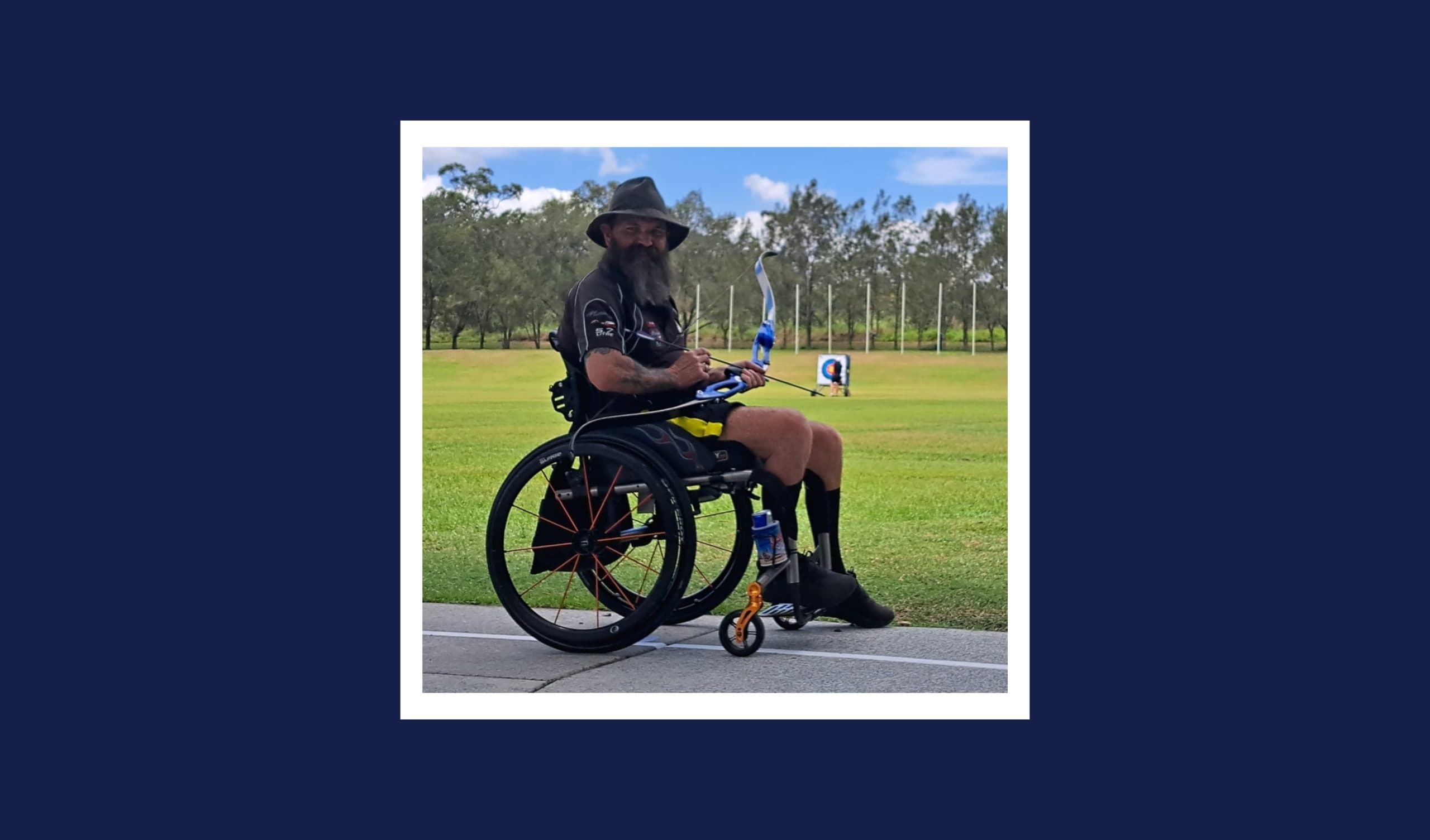 A man with a beard and hat in a wheel chair holds a bow and arrow. Behind him, archery targets, grass and trees are visible.