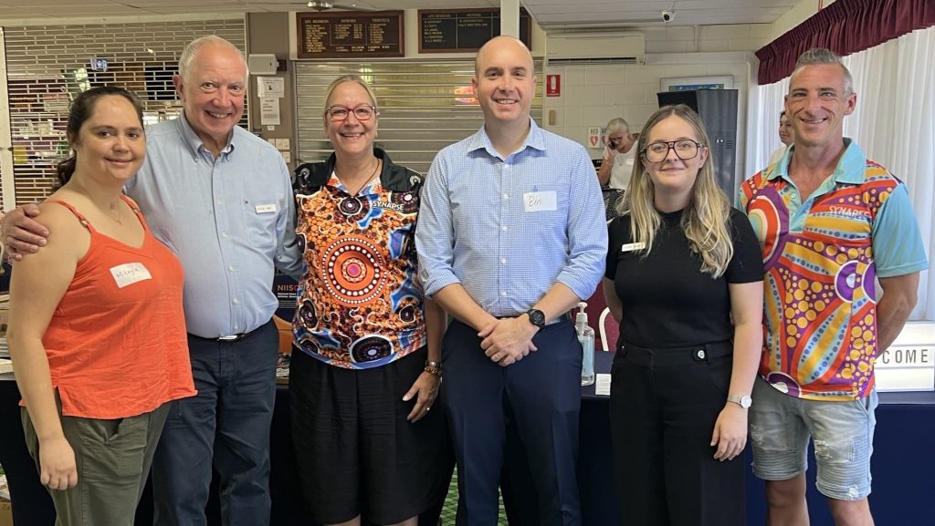 Three men and three women stand in a row smiling at the camera. They are standing on green carpet with bowls club plaques visible in the background.