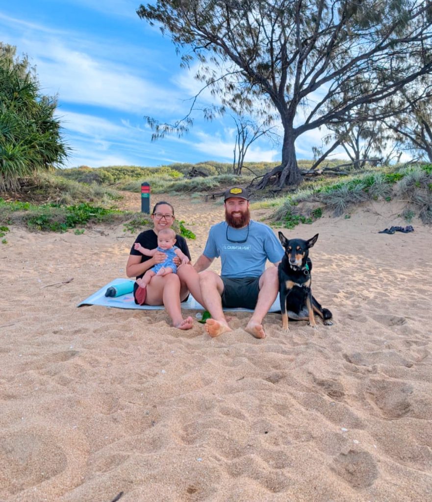 A man, woman and baby sitting on a beach with a dog.