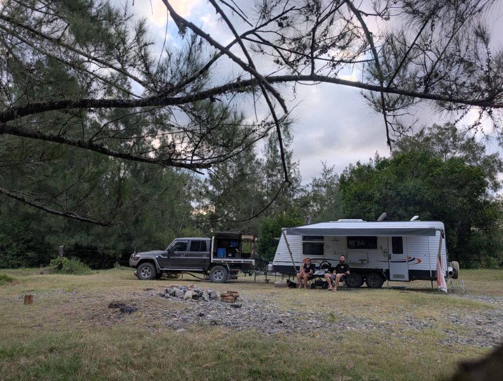A truck and a trailer in a field. Trees are visible above. Two people are sitting in front of the caravan.