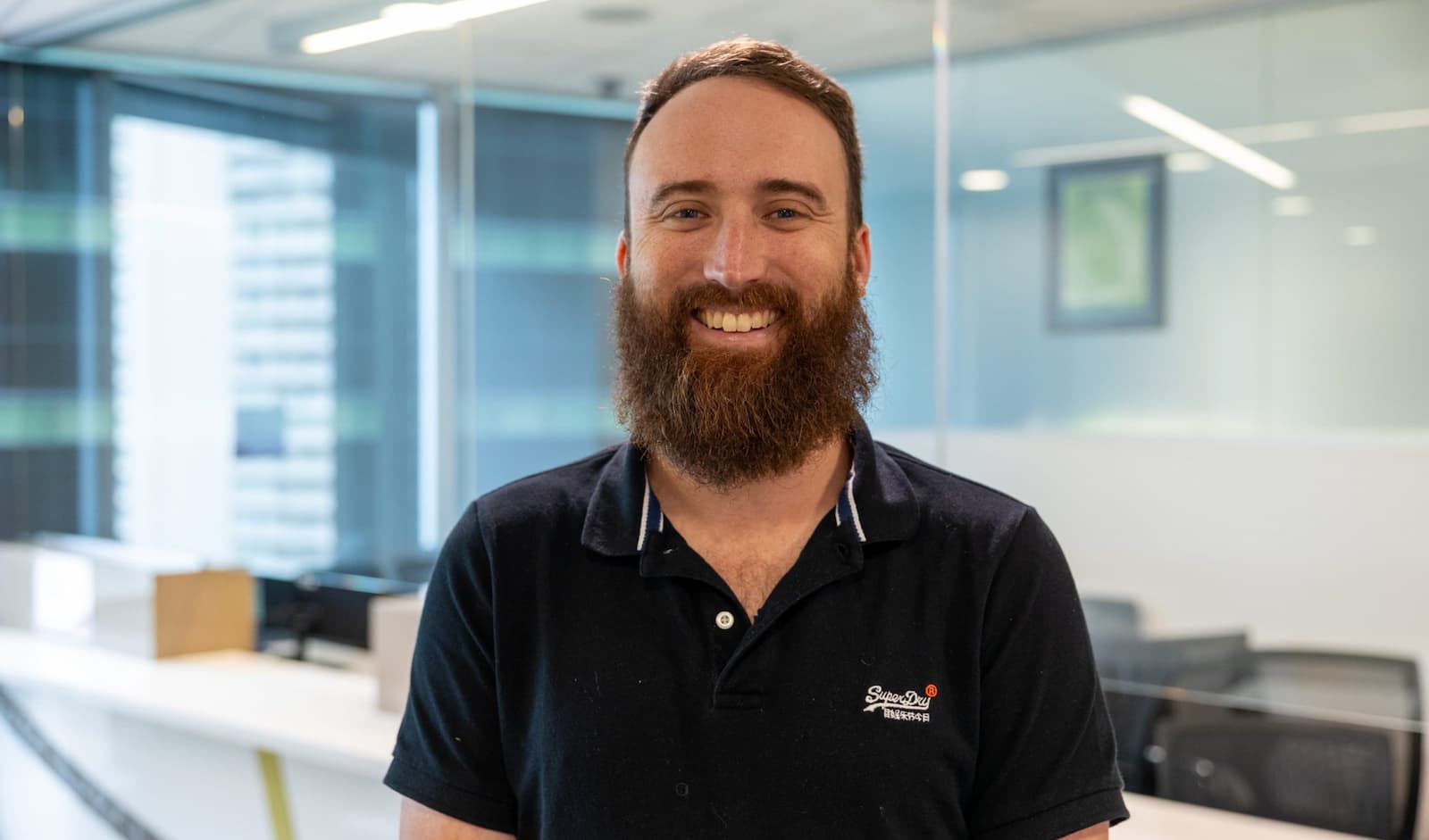 A smiling, bearded man is wearing a black polo shirt as he looks at the camera. Behind him glass, lighting, desks and chairs of an office space are visible.