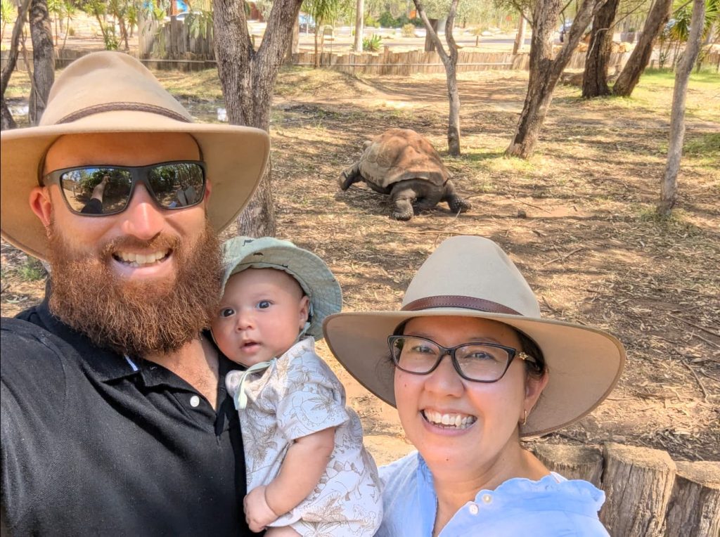 A man and woman holding a baby. A giant tortoise is visible in the background