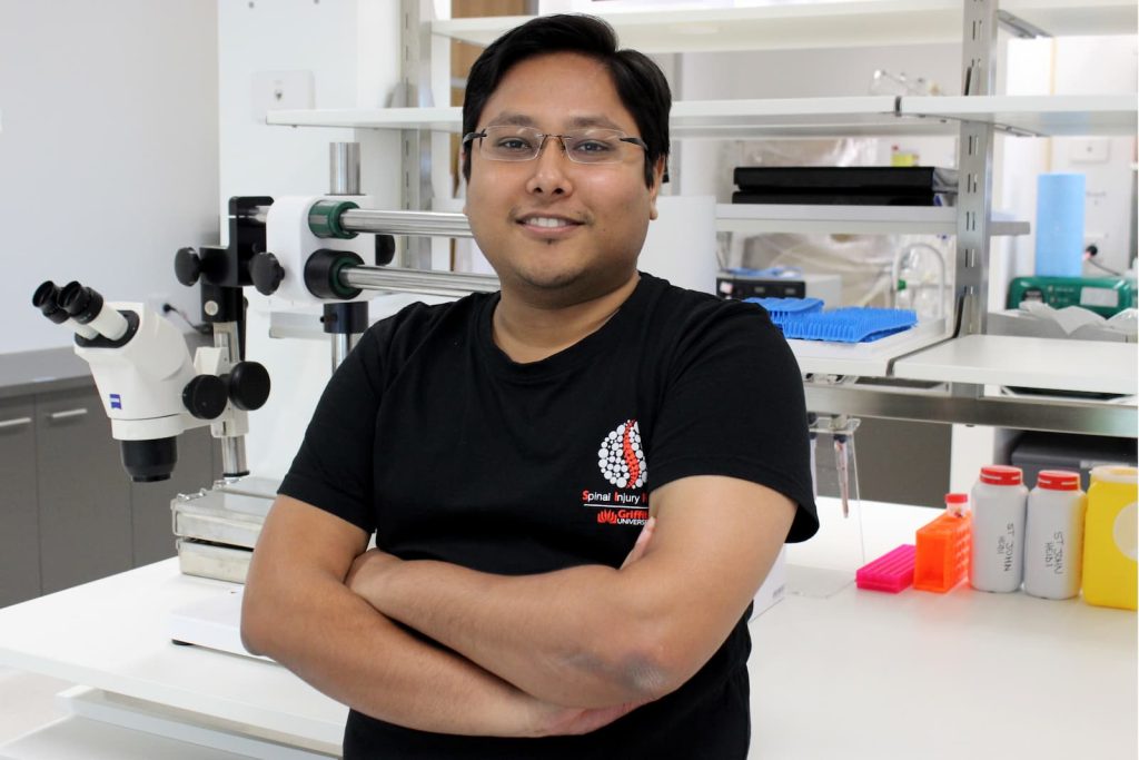 A man (Dr Ronak Reshamwala) wearing a black t-shirt and wire framed glasses stands with his arms folded and smiling in front on laboratory equipment.