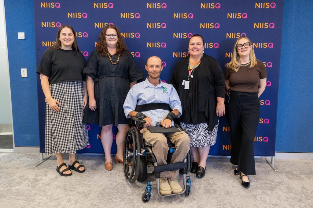 Five people in a row are smiling â A man in a wheelchair is in the middle with two women standing either side of him. Behind them is a navy media wall with âNIISQâ in orange and fuchsia text.