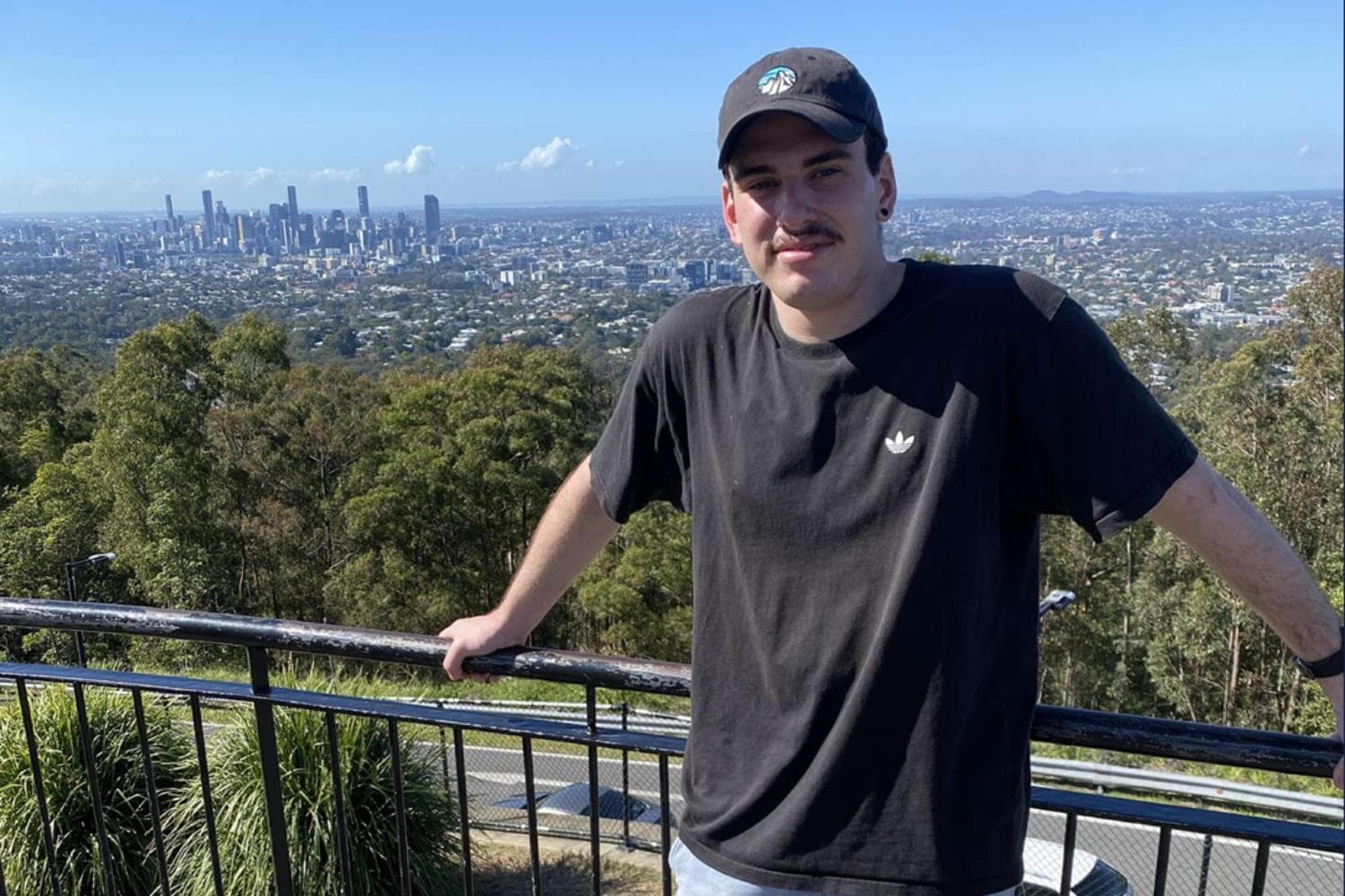 An image of a young man standing at a lookout point with a city skyline and dense greenery in the background under a clear blue sky. He wears a black t-shirt and a dark cap, leaning on a black metal railing overlooking an urban and natural landscape.