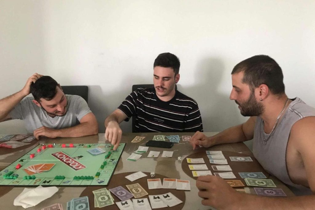 An image of three young men playing a Monopoly board game at a table, with various game cards, money, and property deeds spread out. 
