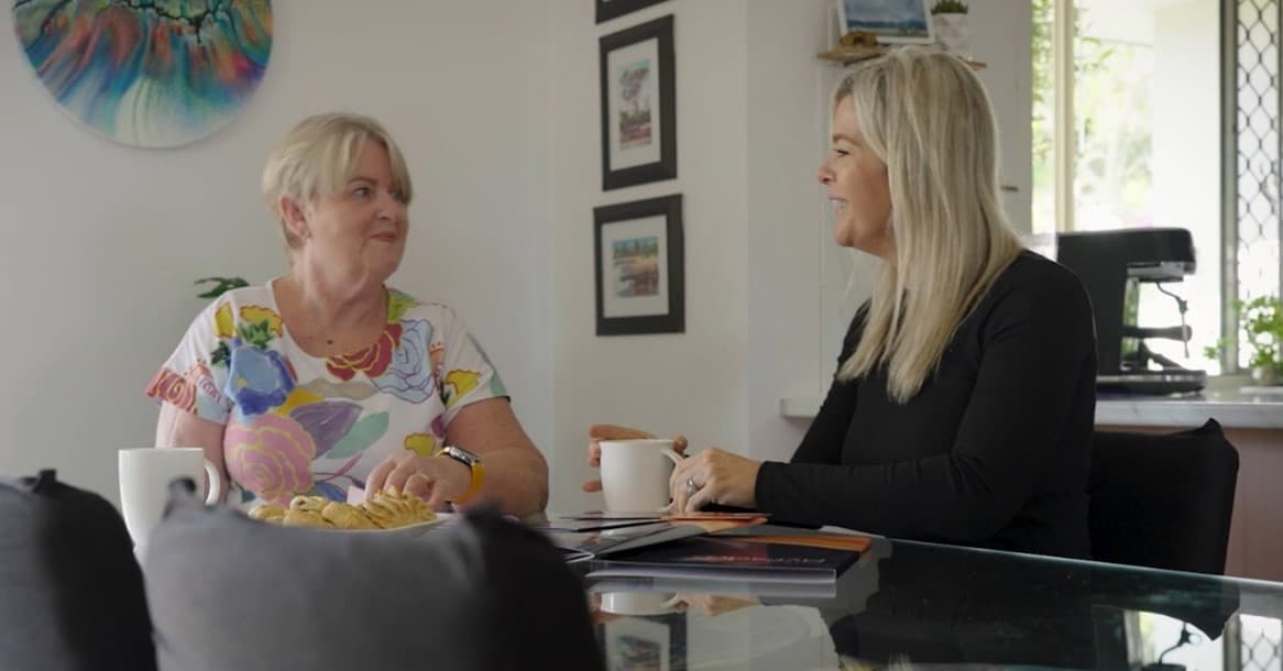 A woman with short blond hair sits across the table from a woman with long blonde hair. They are both smiling and have coffee mugs in front of them.
