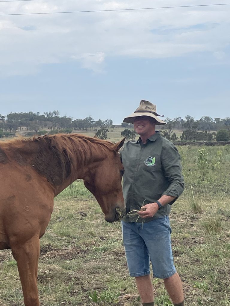 Image of a man smiling at the camera while feeding grass to a brown horse
