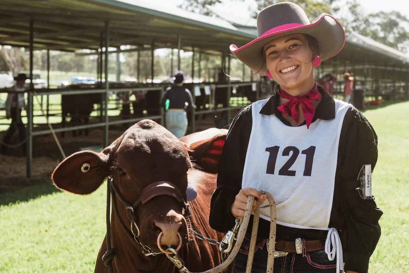 Photograph of a young woman wearing a cowboy hat, numbered vest, and neckerchief standing next to a brown cow with a halter in an outdoor livestock show setting.