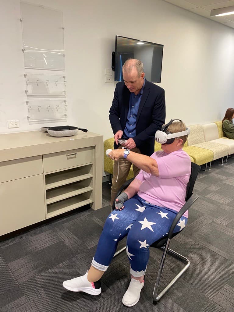 A woman (NIISQ participant) sits in a chair wearing virtual reality goggles, a pink t-shirt and jeans. She holds one arm out straight and holds a white device. A man (Professor Trevor Russell) wearing a navy suit jacket and camel coloured pants stands behind her watching.