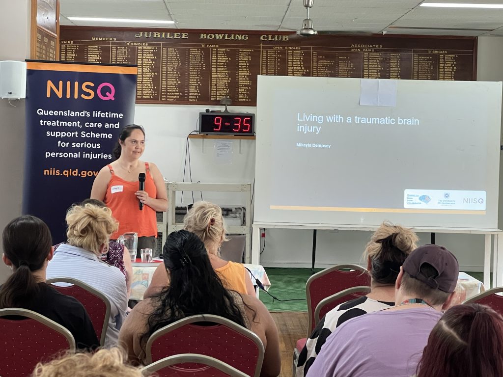 Woman holding microphone addresses room of seated people. She stands in front of a "NIISQ" banner and a PowerPoint presentation that reads "Living with a traumatic brain injury Mikayla Dempsey".