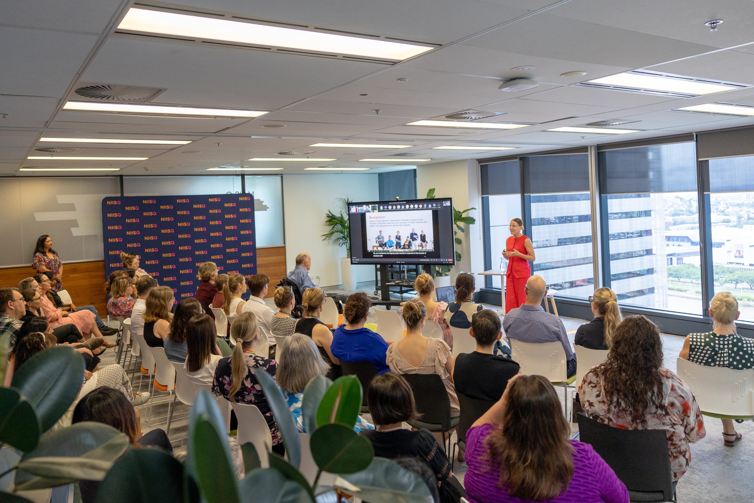 A room of seated people watch as a standing woman wearing all red talks next to a large screen.