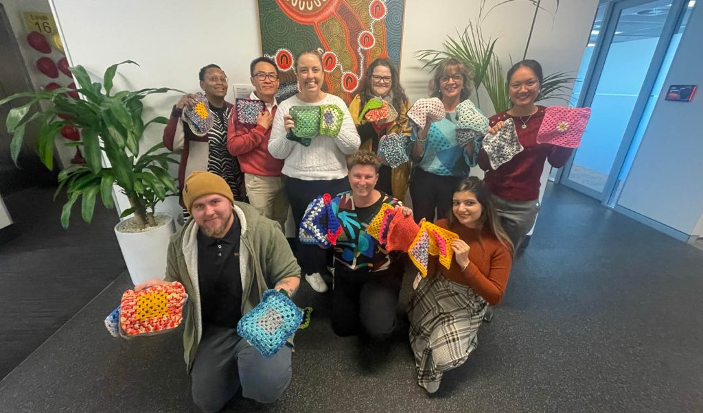 A group of 9, smiling men and women hold up brightly coloured crocheted squares.