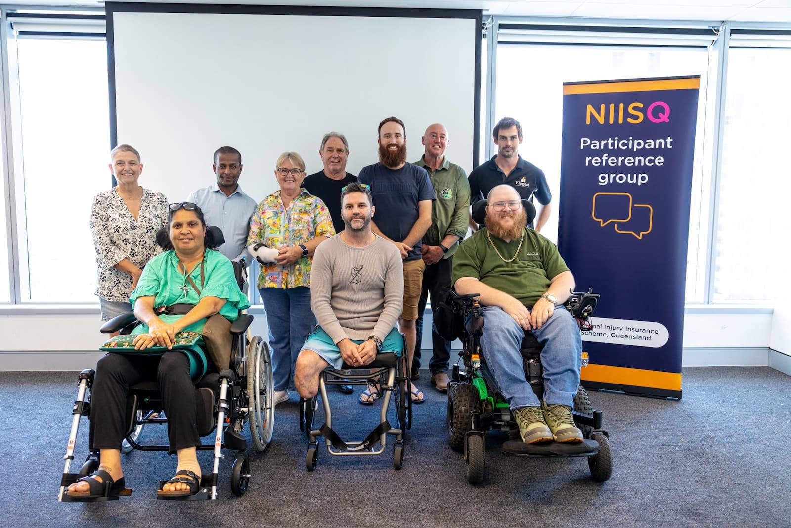 Ten smiling people are together in a group. Three people are in wheel chairs with seven standing behind them. To their left is a banner that reads, “NIISQ Participant reference group.”