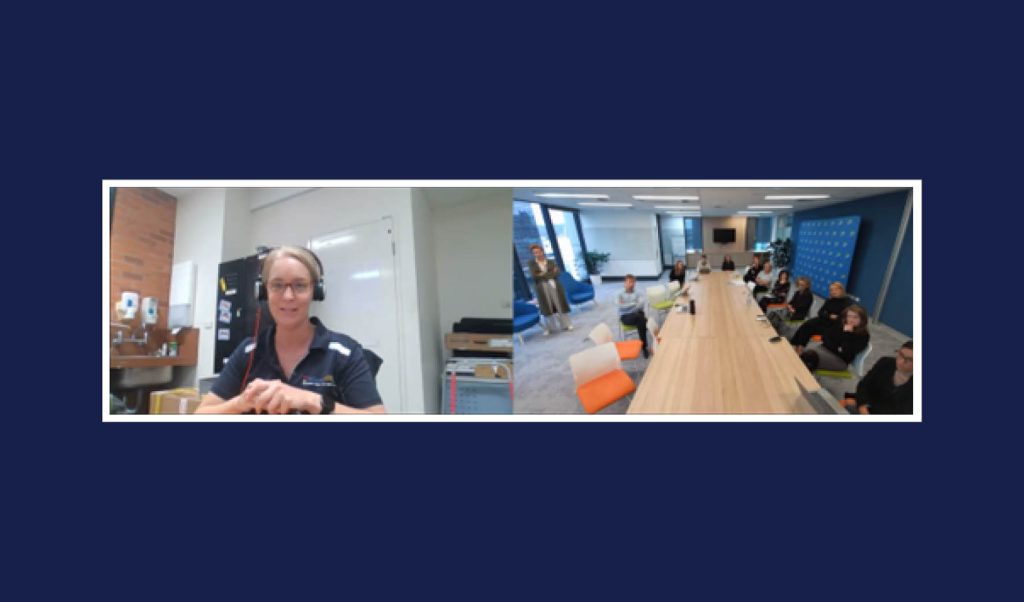 Video call between blonde woman wearing glasses and headphones and a room full of 12 people sitting at a long table.