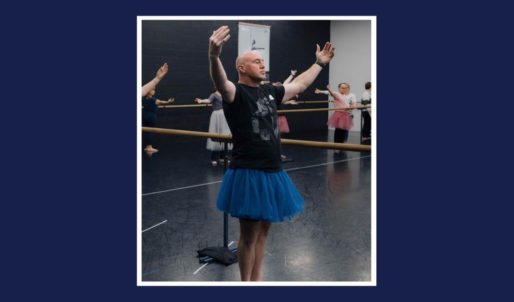 Man wearing black shirt and blue tutu stands with hands raised, fingers facing in, in a ballet position.