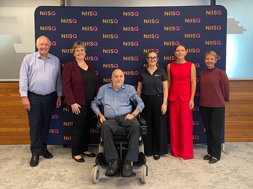 Four women and one man are standing. Another man is in a wheelchair. All are smiling in front of a navy media wall with orange and pink text reading “NIISQ”.