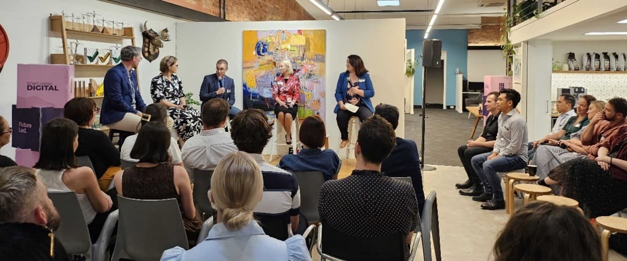 A room full of seated people are listening to two men and three women seated on stools.