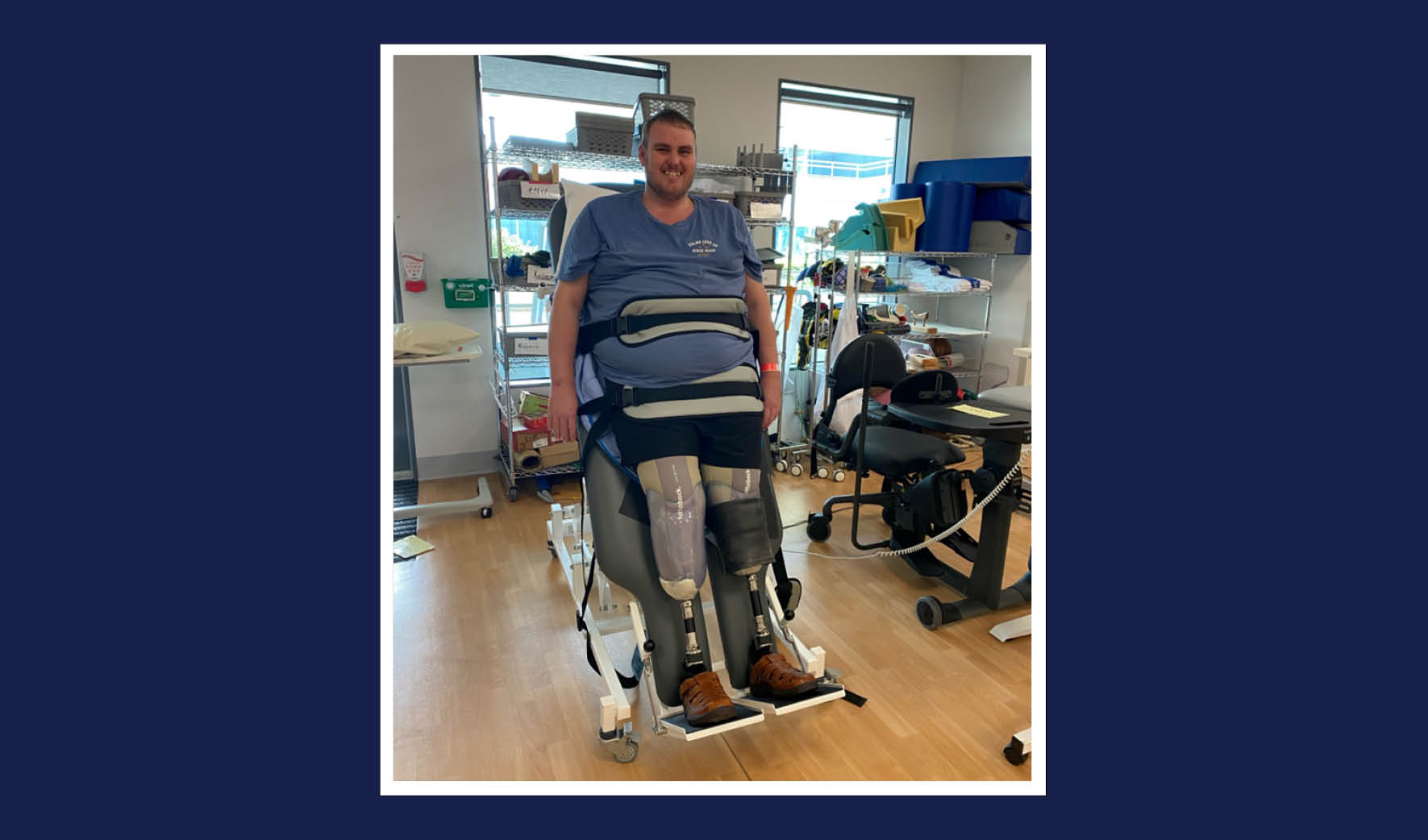 Man with two prosthetic legs and blue t-shirt is smiling. Medical equipment and shelving is visible in the background.