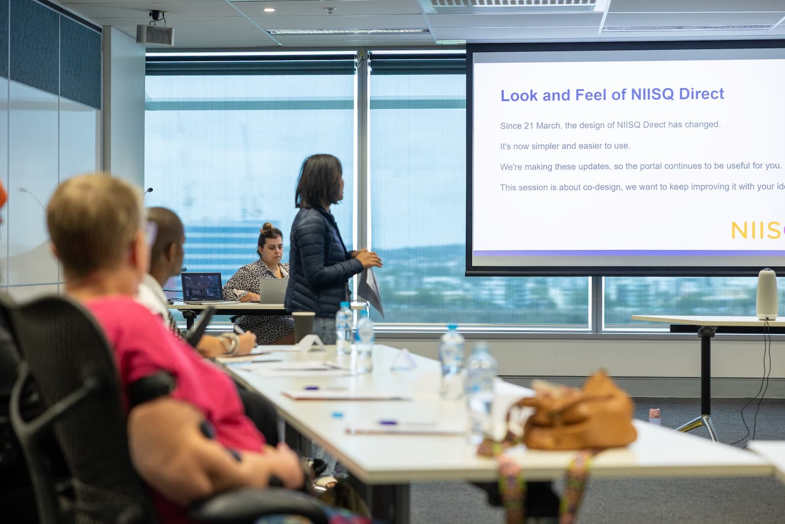 A woman with short, dark hair stands in front of a room full of people. To her right is a large screen that reads, “Look and feel of NIISQ Direct.”