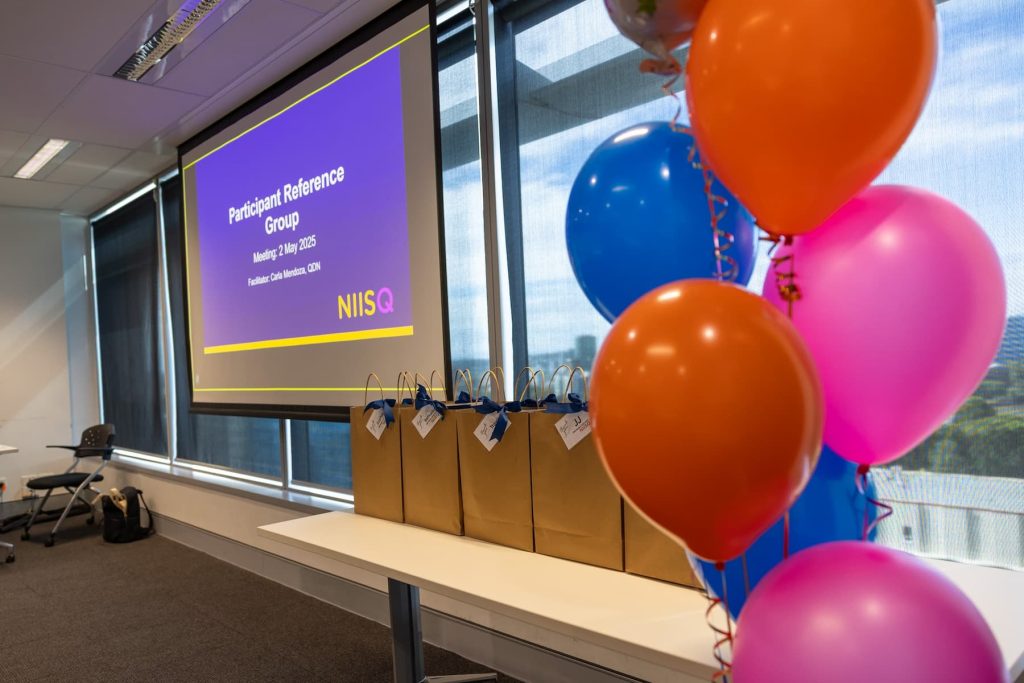 Orange, pink and blue balloons in the foreground. A blue screen with white text that reads “Participant Reference Group” is visible in the background.