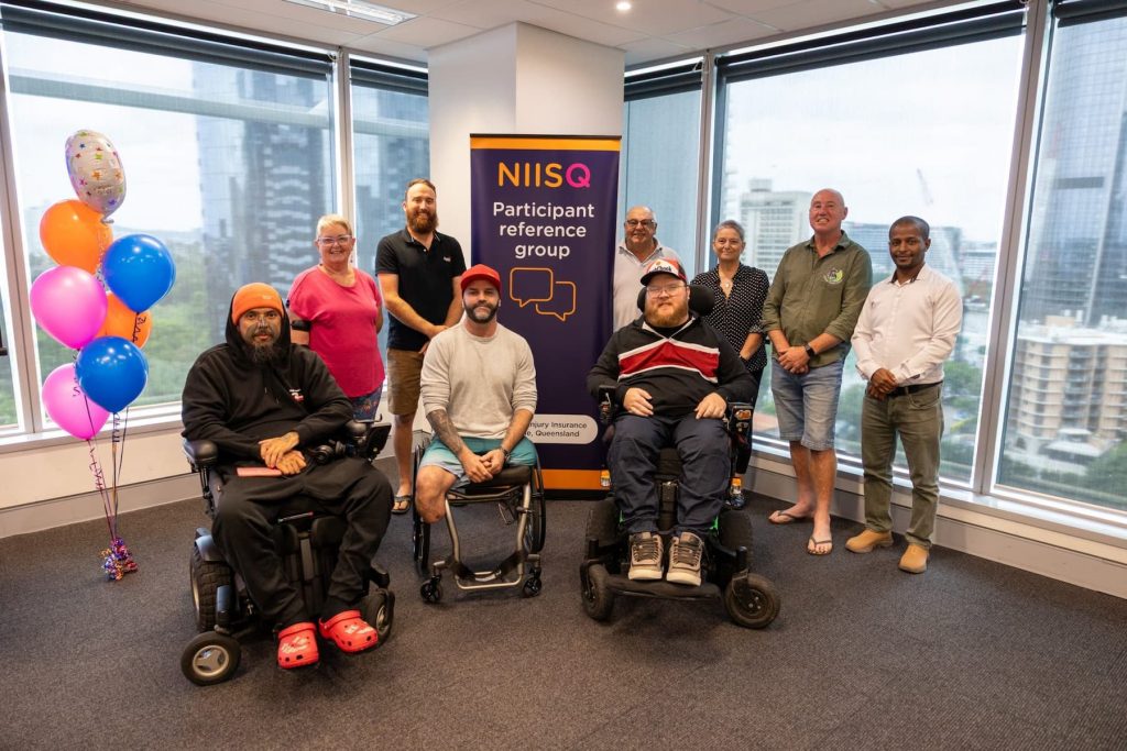 Nine people are smiling in front of a navy, orange, white and fuchsia banner that says “NIISQ Participant Reference Group”. Three men in wheelchairs are in the foreground. Two women and four men stand at the back. A city scape is visible through windows behind them. To the left is a bunch of pink, orange and blue balloons.