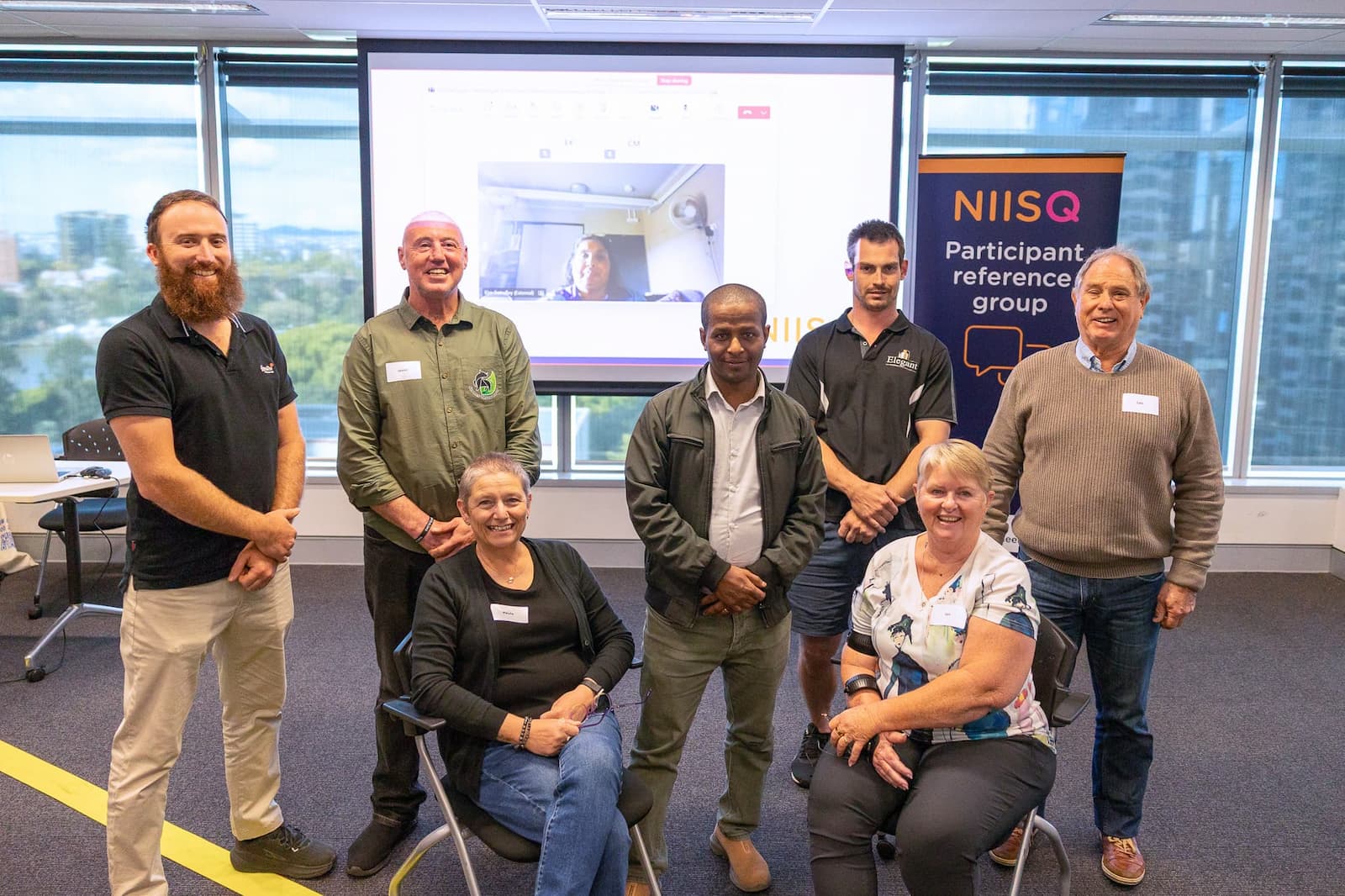 Seven people in a group smiling. Two women are seated at the front. Five men stand at the back. Behind them, a woman’s face is also visible on a large screen. To the left of the group is a navy, orange, pink and white banner that reads “NIISQ Participant reference group”.