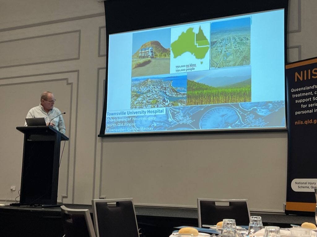 Man stands at podium on stage looking towards a PowerPoint presentation. The presentation includes aerial photos of landscapes, an image of a truck, a cane field and a map of Australia.