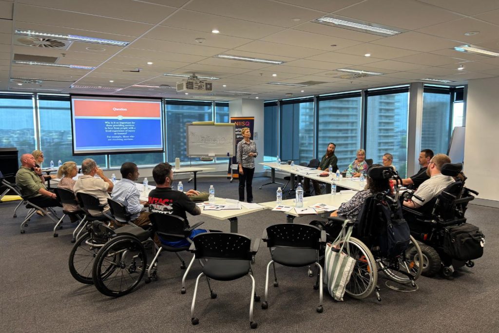 An image of a diverse group of people, including individuals in wheelchairs, attending a presentation in a modern conference room with large windows and city views. A female presenter stands near a whiteboard and a screen displaying a slide titled "Question," while attendees sit around U-shaped tables with water bottles and paper.