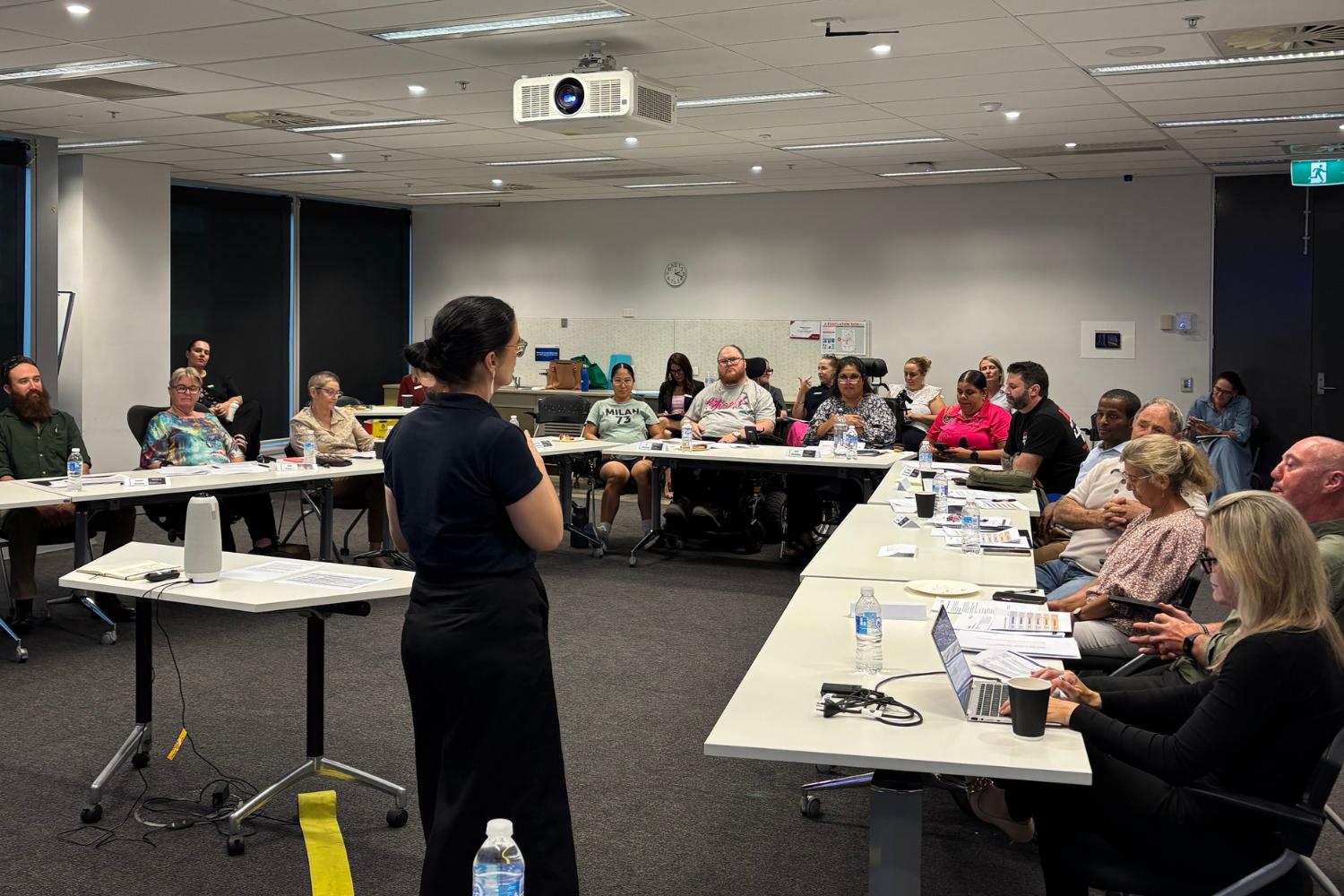 An image of a meeting or workshop showing a female presenter standing and addressing a diverse group seated around U-shaped tables in a conference room.