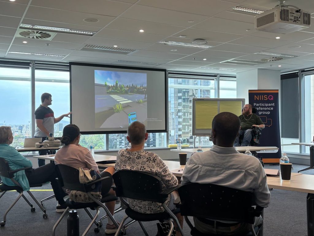 Two men are presenting to a room of seated people. One man stands, talks and gestures to a large screen that includes a rendered road scape. The other, in a wheelchair watches the screen.