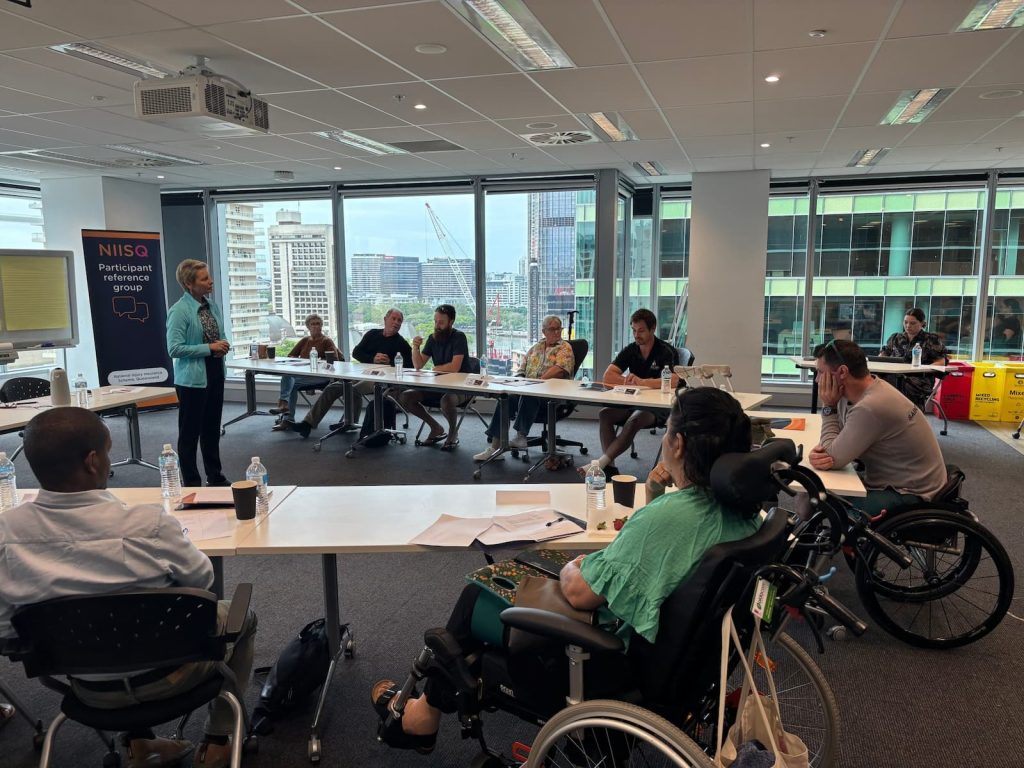 Standing woman with short blonde hair talks to people at desks. A city scape is visible through windows behind them.