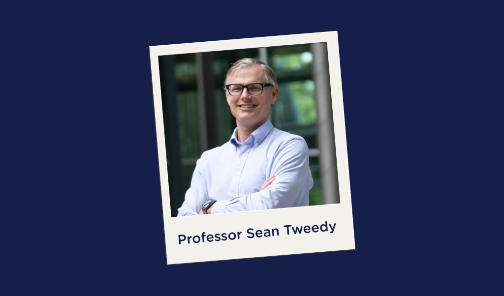 A polaroid picture on a navy background of a man wearing glasses and a light blue button-up shirt smiling with his arms crossed. Behind him greenery is visible through windows. On the polaroid picture there is navy text that reads “Professor Sean Tweedy.”