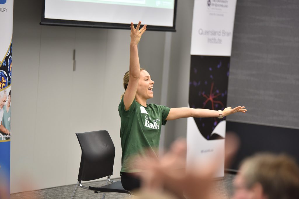 A blonde woman holds her arms - one straight up, the other out. She wears a green t-shirt and is seated. Others’ hands are visible in the foreground.
