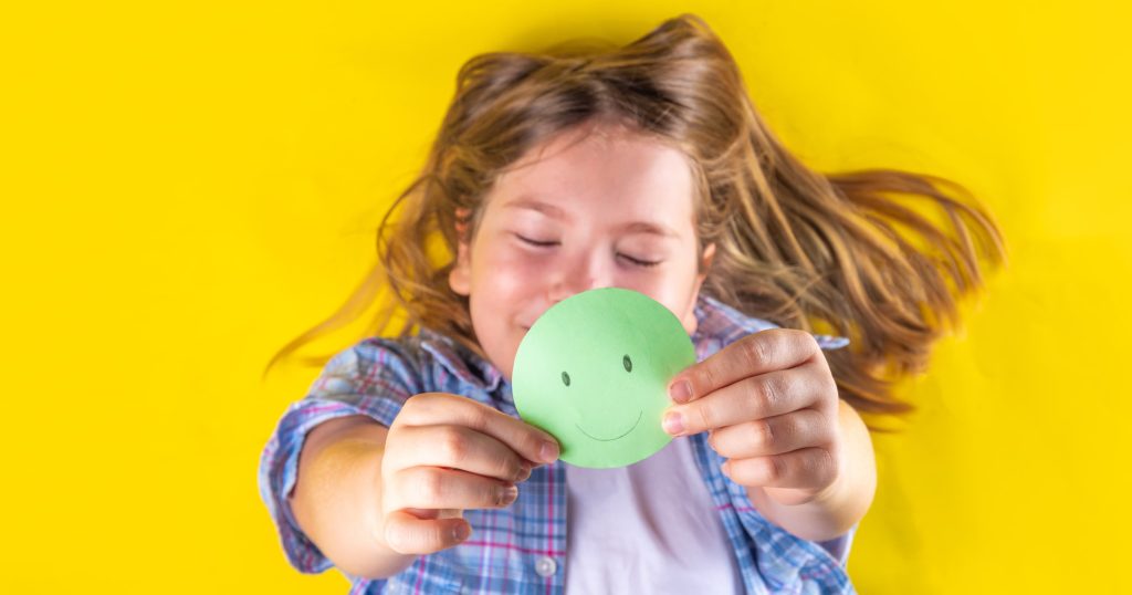 Girl holds up green smiley face cut out of green paper. The background is yellow.