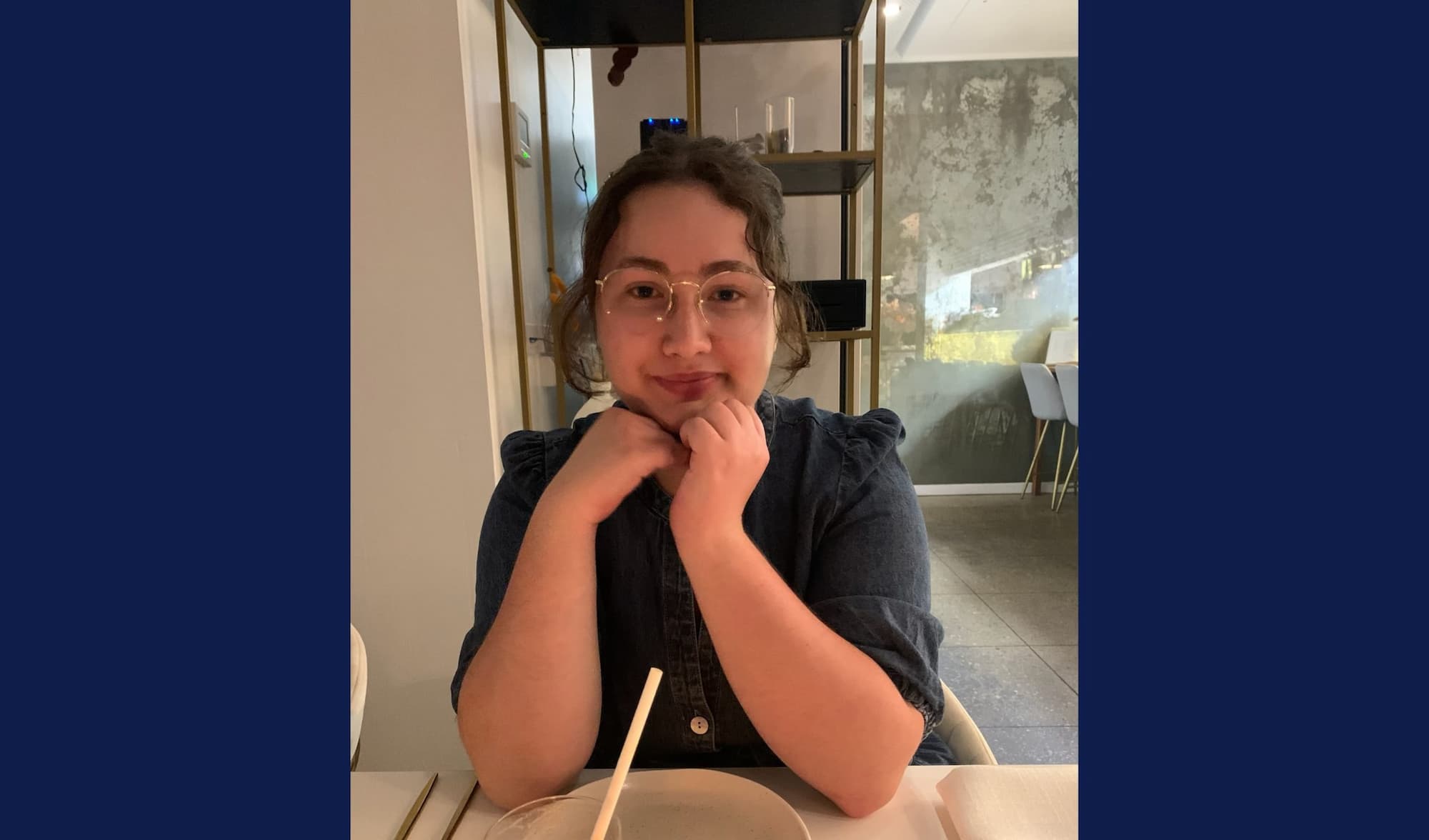 A woman with dark, curly hair, wearing a navy button-up top and glasses sits at a table -- lightly smiling. Navy strips are visible either side of the image.