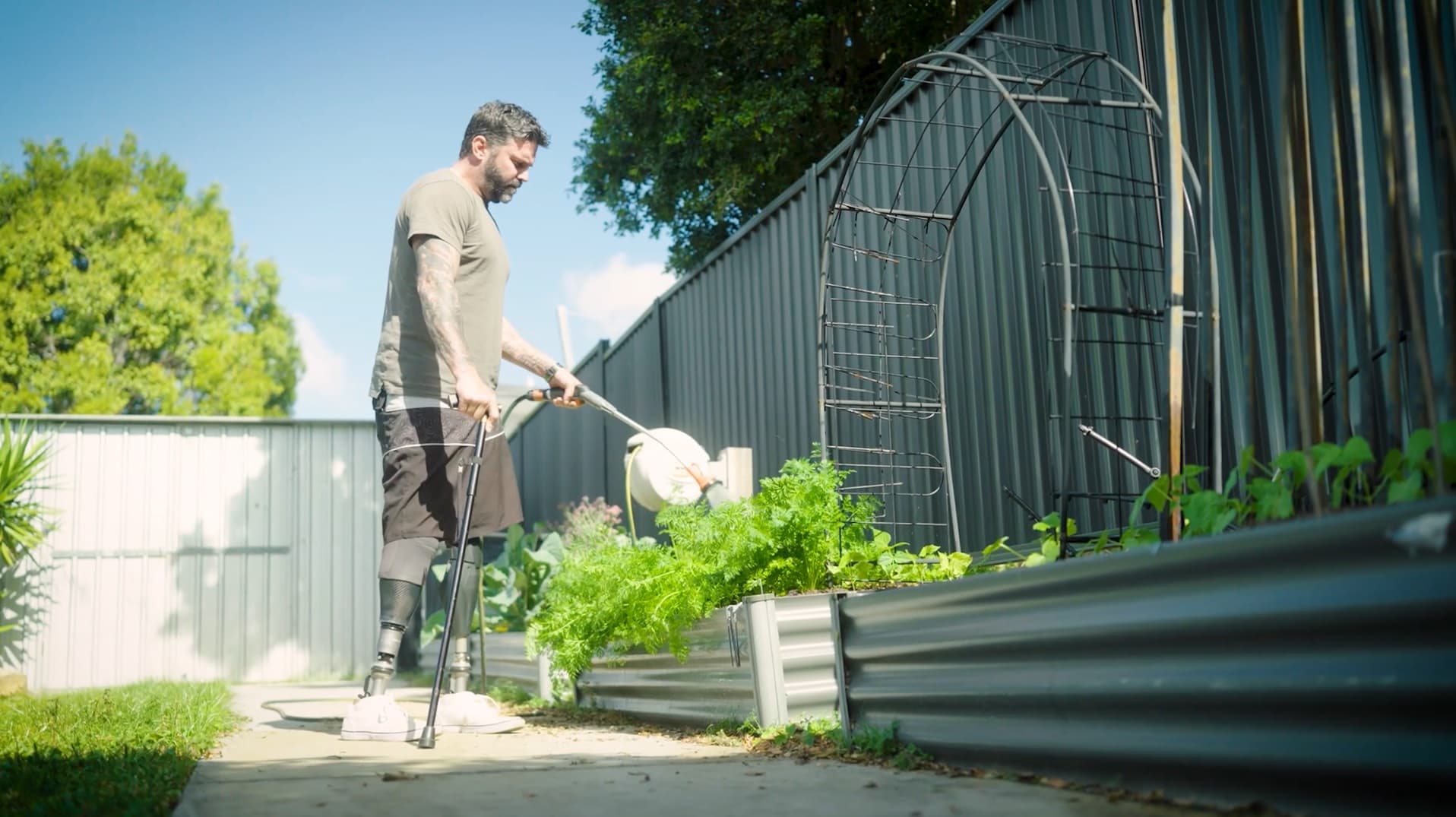 A man with prosthetic legs wearing neutral-coloured shorts and shirt waters a garden.