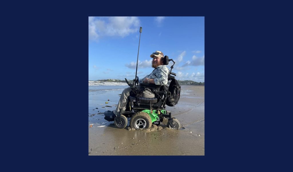 A smiling, bearded man is in a wheelchair on a beach. Blue sky, sand and water are visible behind him.