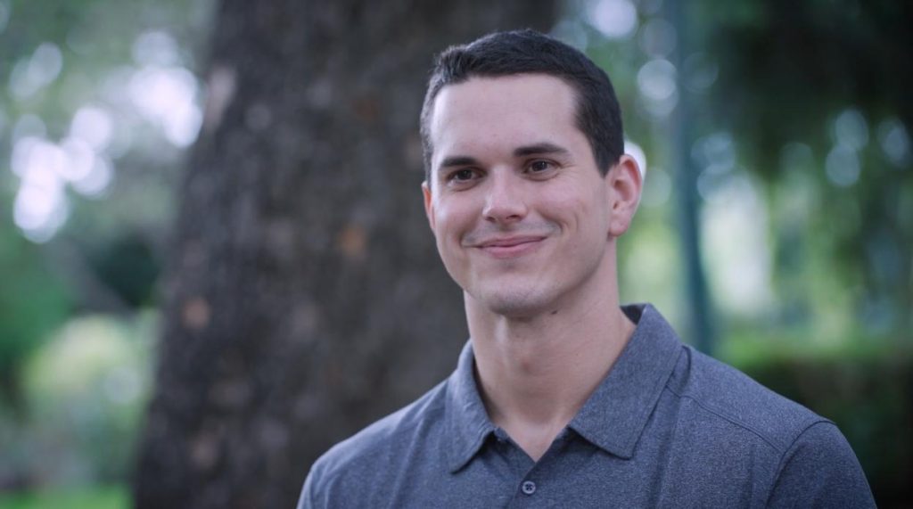 Clos up of a man wearing a grey shirt, smiling. Trees are visible behind him.